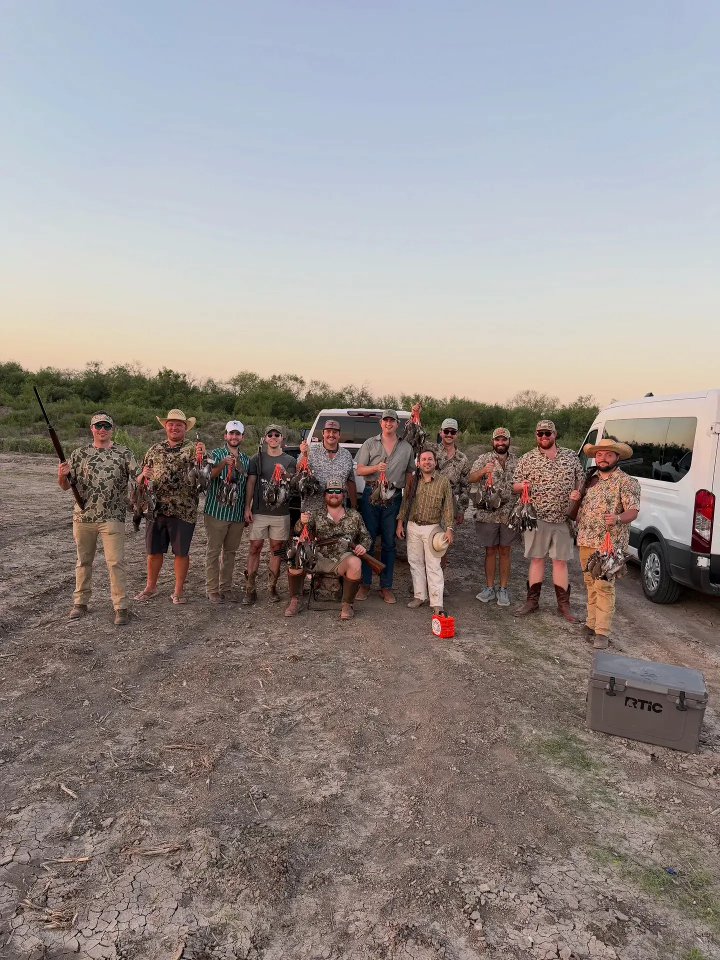 A group of hunters posing with shotguns in a dirt lot at sunrise